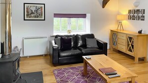 A living room with a black leather sofa, a wooden coffee table, a radiator, a window with a view of greenery, and a framed picture on the wall. - 3 bedroom accommodation in Steading, Berwick-upon-Tweed (Steading, Berwick-upon-Tweed)