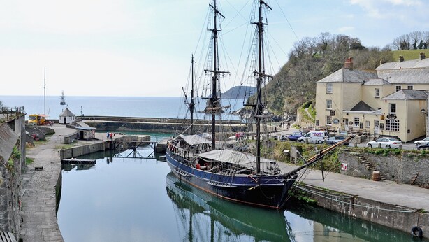 Ett historiskt segelfartyg låg förtöjt vid en hamn, och i närheten fanns en byggnad och en väg.