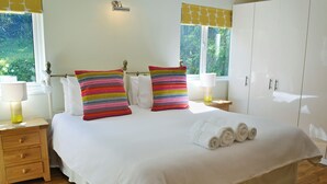 Een keurig opgemaakt bed met witte lakens, twee gestreepte kussens en opgerolde handdoeken op een houten vloer met een kamerkleed.