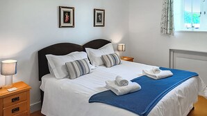 Una cama bien hecha con ropa blanca y un plaid azul, dos lámparas de noche, una mesita de noche de madera con una lámpara, y dos cuadros en la pared.