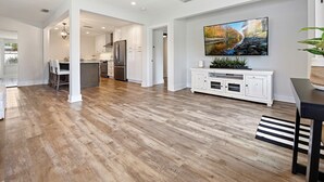 A spacious living area with hardwood flooring, a kitchen in the background, and a large flat-screen TV mounted on the wall. - SaltRunner: Updated 3/2 home with Private Pool (St. Augustine)