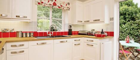A kitchen with white cabinets, a wooden countertop, a red and white leaf-patterned valance, and a view of greenery outside.