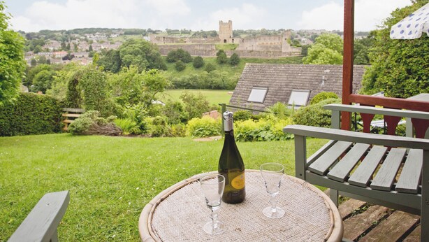A table with a bottle and glasses on a wicker table, overlooking a grassy area with a building in the background.