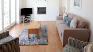 Un salon comprenant un canapé, deux fauteuils à rayures, une table en bois, un téléviseur et des œuvres d’art accrochées aux murs.