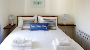 Un lit bien fait, avec des draps blancs et des oreillers à rayures, deux serviettes blanches pliées et un oreiller bleu orné du texte « Soleil, mer, sommeil ».