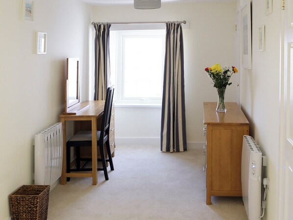 A room with a wooden desk, a chair, a radiator, a window with curtains, a vase with flowers, and a radiator cover.