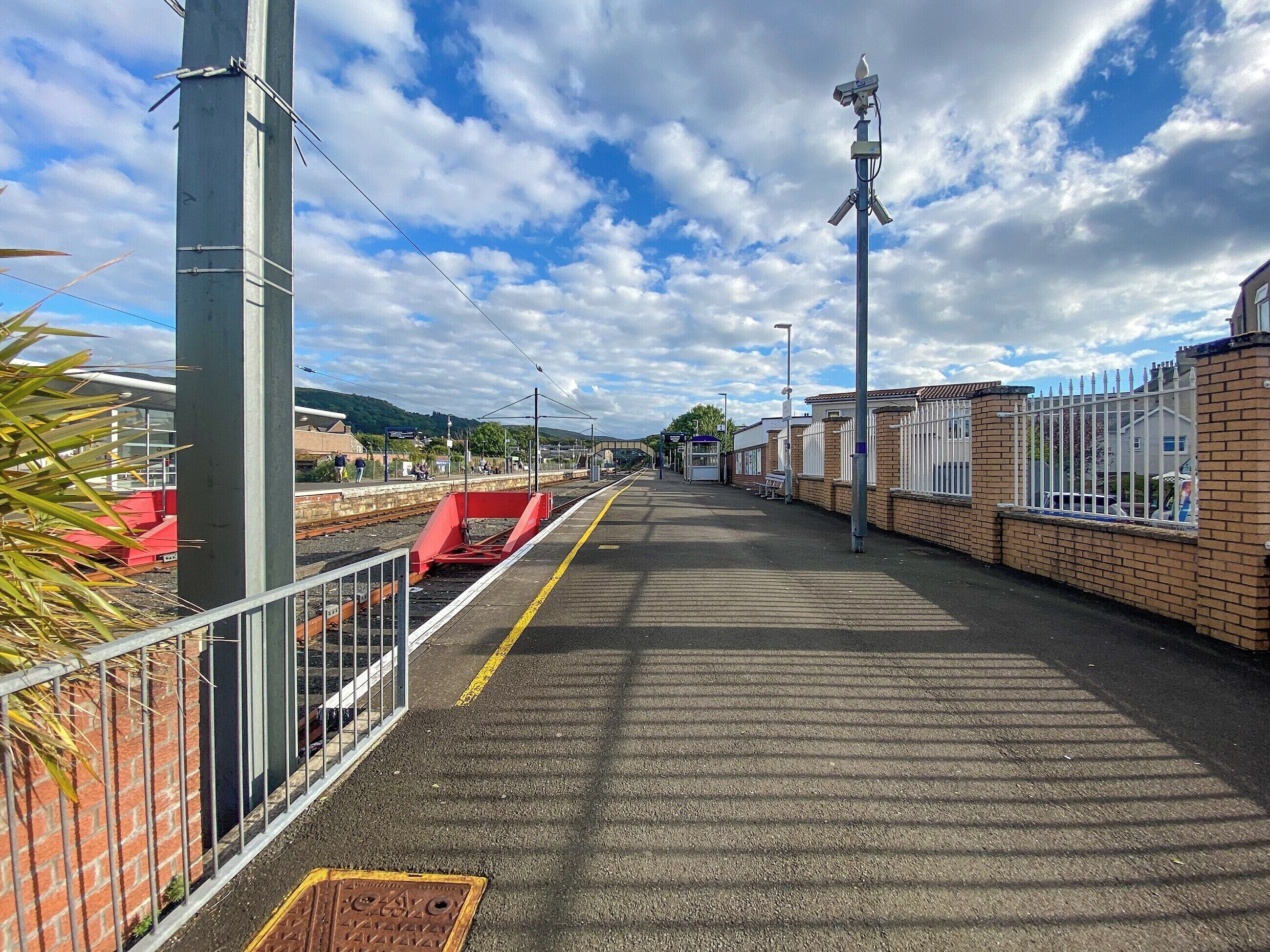A train station platform with a clear sky, a fence, and a pedestrian walkway.