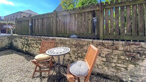 An outdoor seating area with two wooden chairs and a small round table, surrounded by a stone wall and a wooden fence.