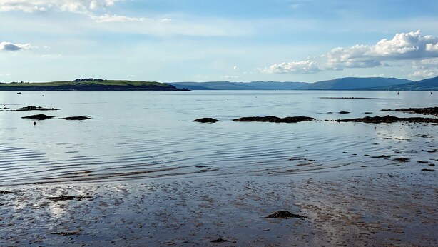 A calm lake with rocky shores and distant hills under a partly cloudy sky.