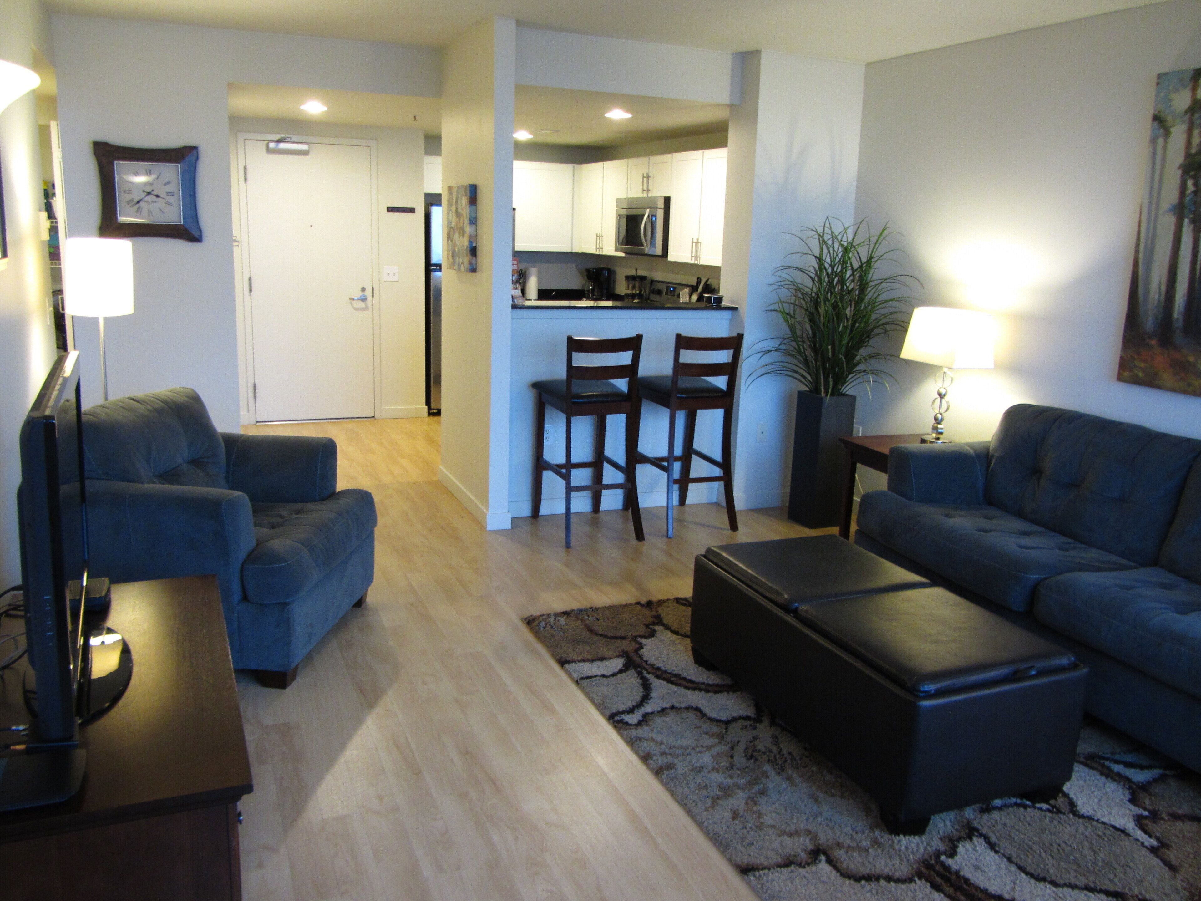 A modern living room with a sofa, two chairs, a coffee table, and a TV. There is a kitchen area with a microwave and a refrigerator.