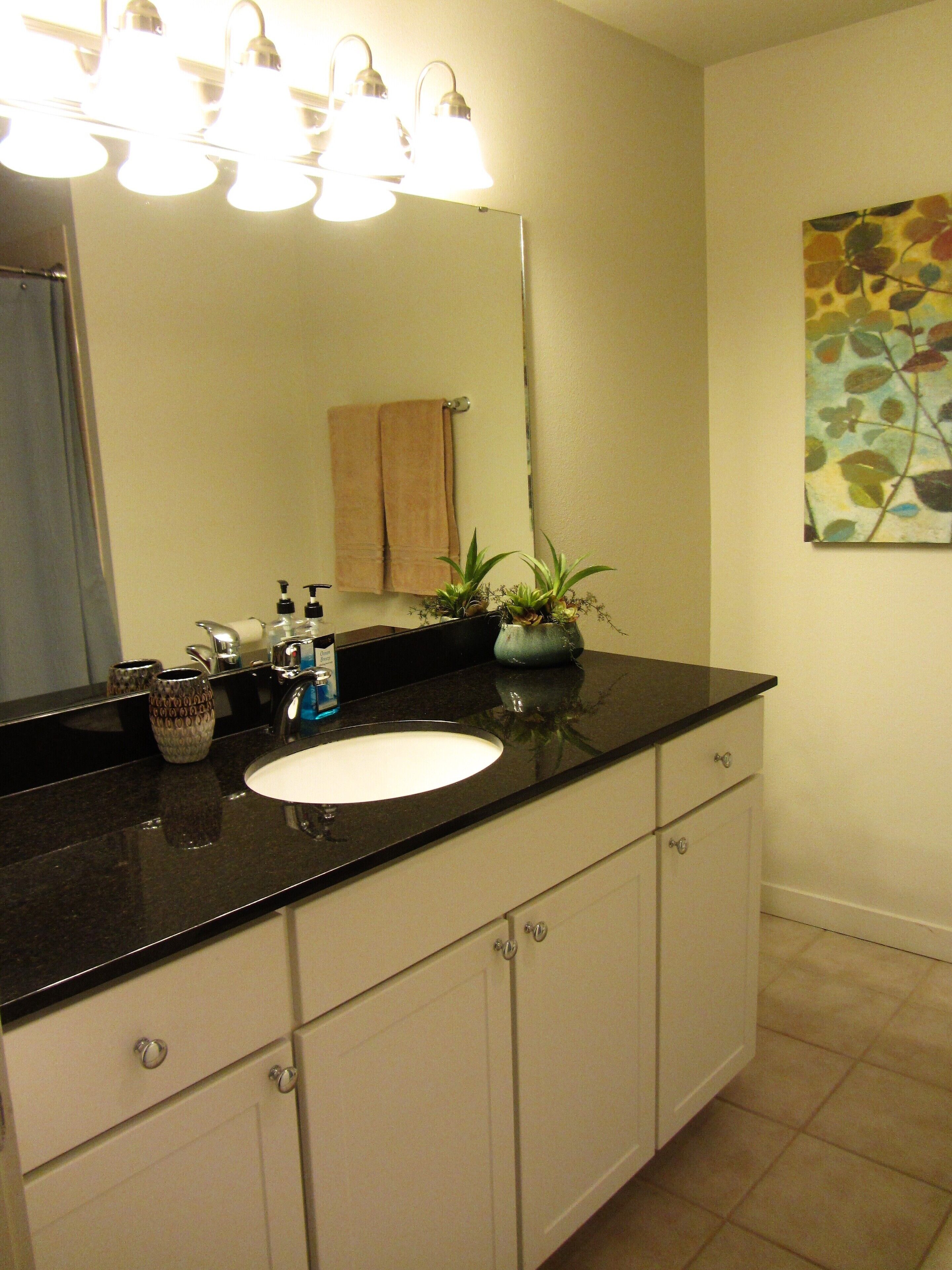 A bathroom with a black countertop sink, white cabinets, and a mirror above it. There are three lights above the mirror, and a towel hangs on the wall.