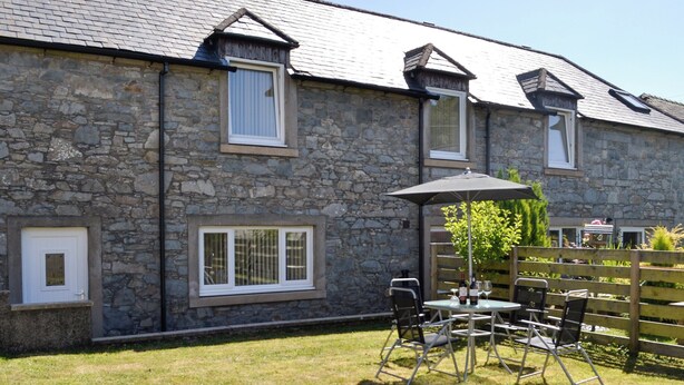 A stone building with a grassy yard, a patio table with chairs, and a wooden fence. - Kirroughtree Steading 1 - UK5859 (Newton Stewart)