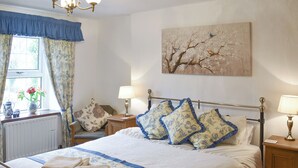 A neatly made bed with floral pillows, a bedside table with a lamp, a radiator, and a window with curtains.