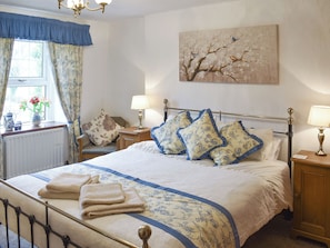 A neatly made bed with floral pillows, a bedside table with a lamp, a radiator, and a window with curtains.