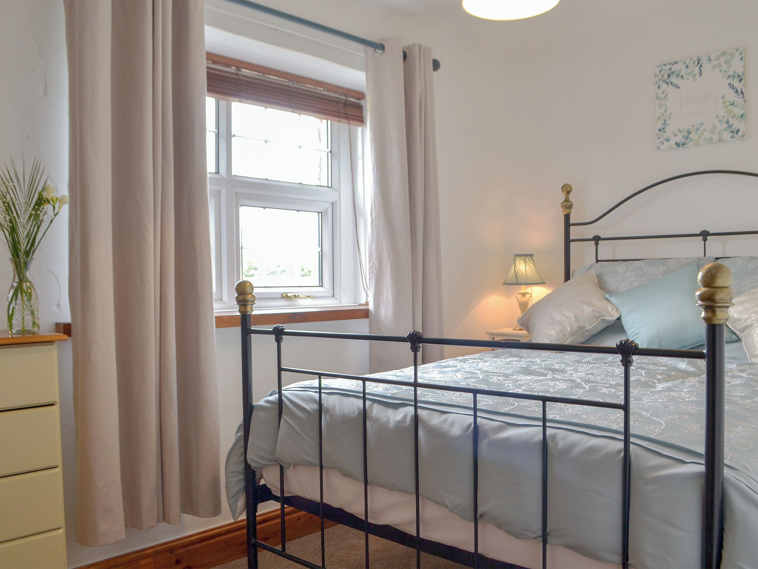 A bedroom with a metal bed frame, a bedside table with a lamp, a window with curtains, and a wall decoration.