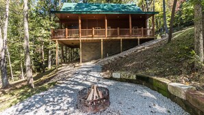 Eine Hütte mit überdachter Veranda, umgeben von Bäumen und einem Kiesweg, der zu einer Feuerstelle führt.