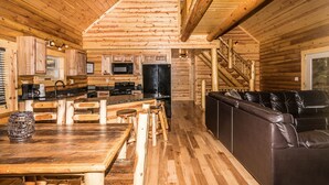 Ein Blockhaus-Interieur mit einem Holzesstisch, Barhocker, einer schwarzen Ledercouch, einer Küche mit Geräten und einer Treppe.