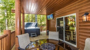 Eine Holzterrasse mit Whirlpool, Glastisch und Stühlen mit Blick auf eine bewaldete Landschaft.