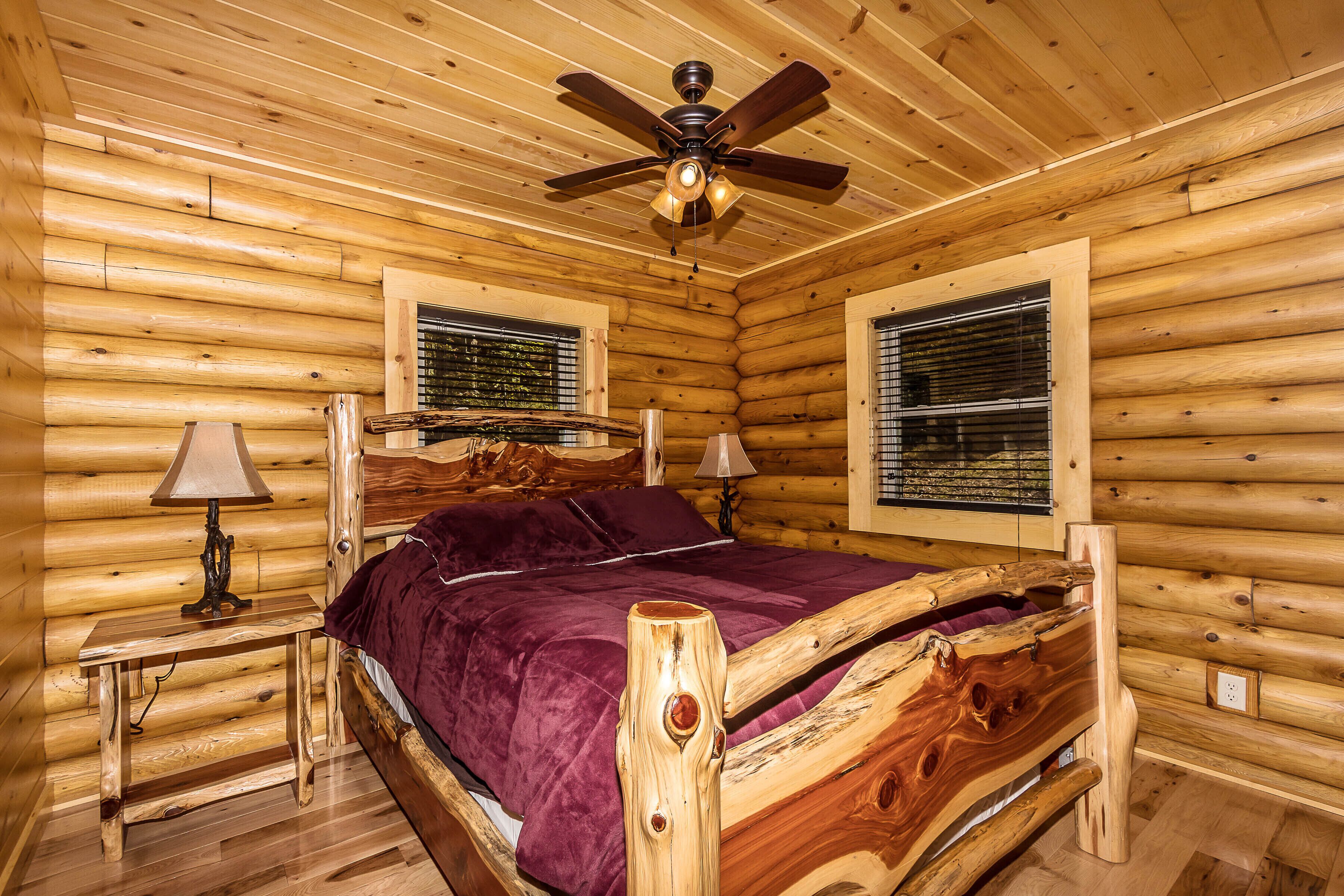 Ein Schlafzimmer in einer Blockhütte mit einem Holzbett, zwei Nachttischen mit Lampen, einem Deckenventilator und zwei Fenstern mit Jalousien.