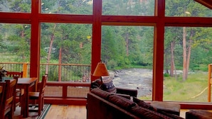 Een kamer met grote ramen met uitzicht op een bos en een rivier. Er staat een houten bank en een tafel met stoelen.