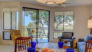A spacious living area with a large sliding glass door leading to a balcony, a comfortable sofa, a dining table with blue glassware, and a small decorative vehicle on the table. - Spacious End Unit (Kihei)