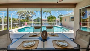 Comedor con una mesa preparada para cuatro, con vista a una piscina, palmeras y cielo despejado.