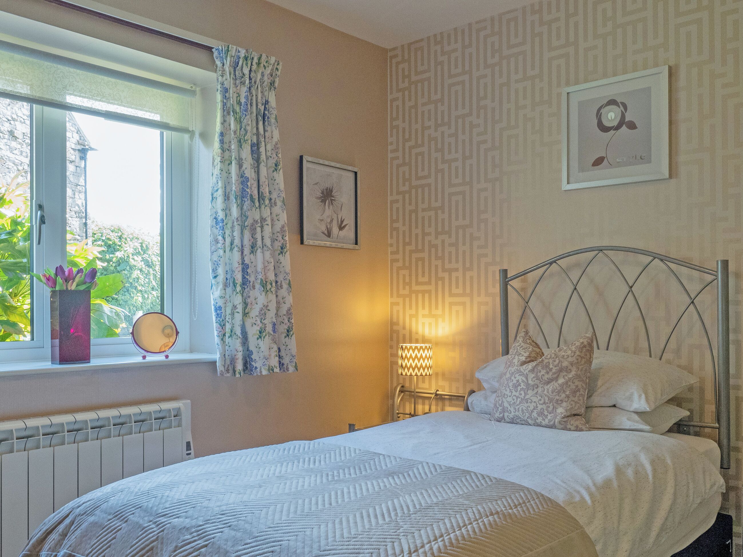 A bedroom with a bed, a window with curtains, a radiator, a wall with patterned wallpaper, and a framed picture.