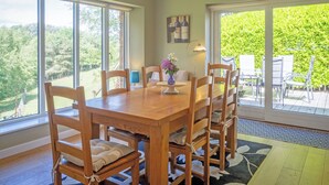 Een eetgedeelte met een houten tafel en stoelen, grote ramen en uitzicht op groen.