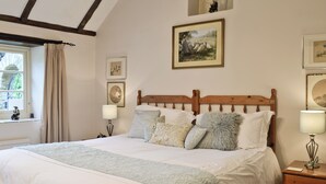 A bedroom with a large bed, wooden headboard, and multiple pillows. There are two bedside tables with lamps, a window with curtains, and framed pictures on the walls. - 1 bedroom accommodation in Ampney Crucis, near Cirencester (Ampney Crucis, near Cirencester)