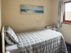 Une chambre avec un lit simple, un tableau représentant une scène de plage, une fenêtre avec des rideaux et une petite figurine posée sur la table de chevet.