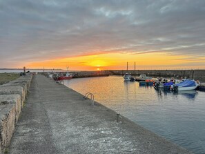 Une scène au bord de l’eau, avec des bateaux amarrés à un quai, une promenade en béton et un coucher de soleil en arrière-plan.