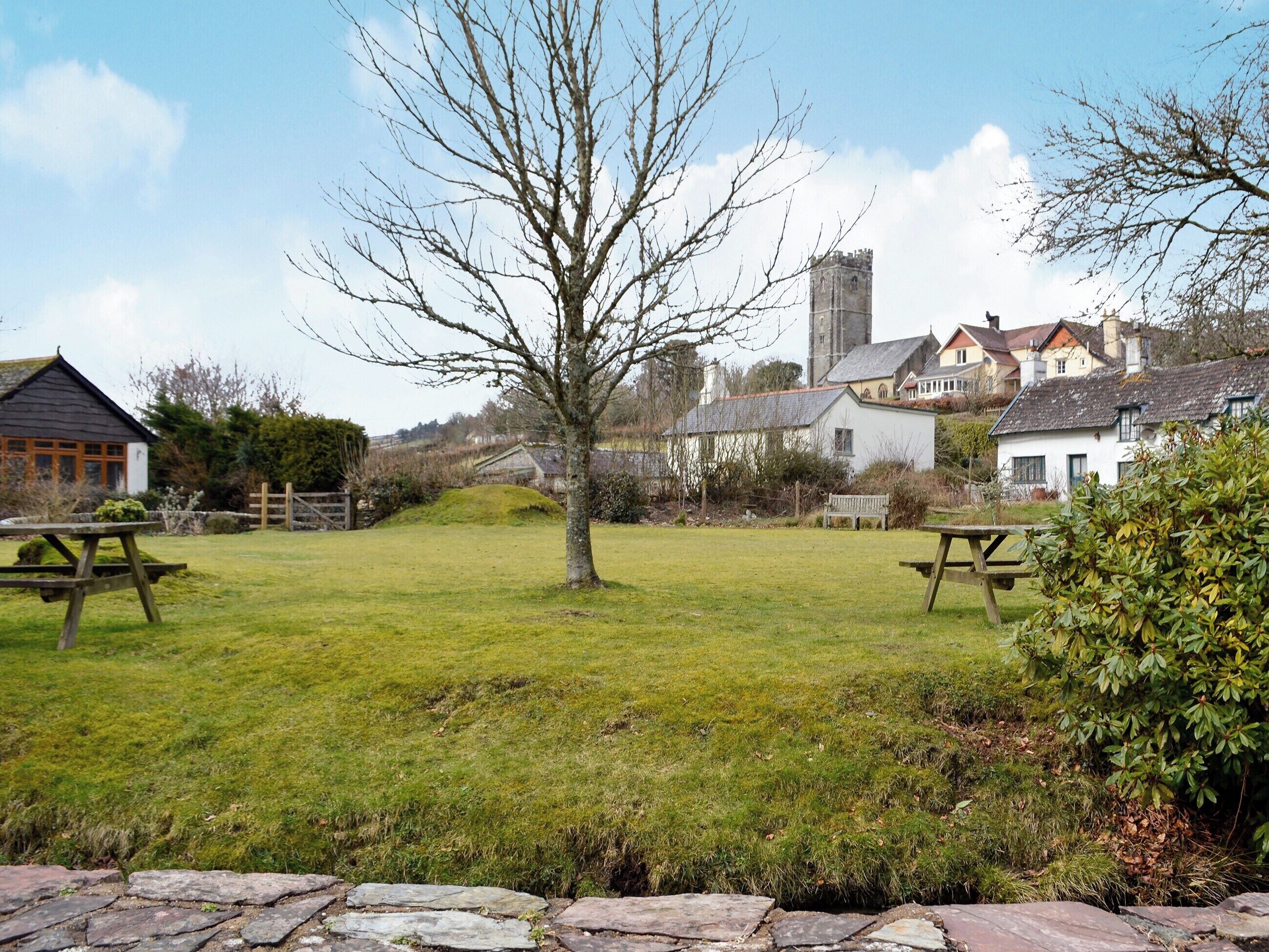 A grassy area with picnic tables, a stone wall, and houses in the background.