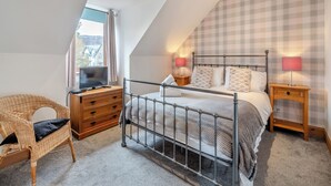 Een slaapkamer met een schuine kap, een bed met metalen frame, twee nachtkastjes met lampen, een houten dressoir, een rieten stoel en een raam met gordijnen.