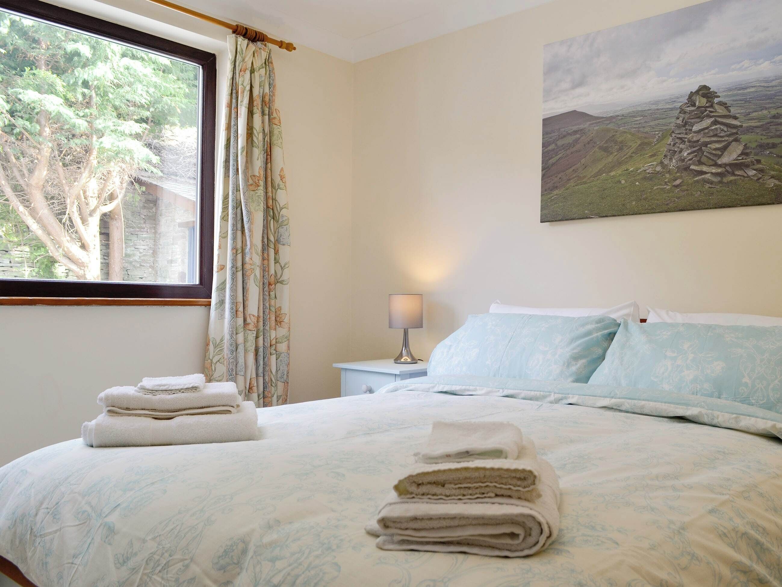 A bedroom with a bed, a nightstand, a lamp, a window with curtains, and a painting on the wall.