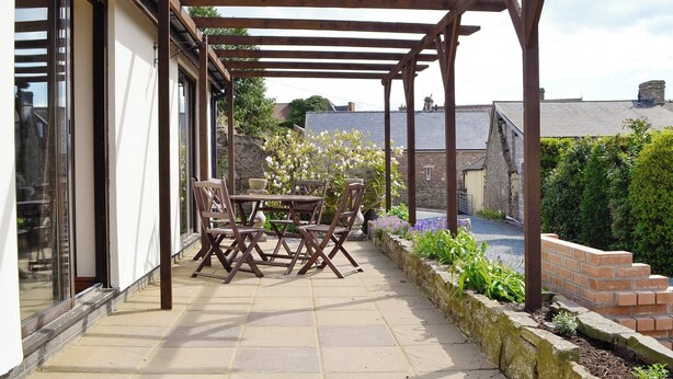 Une terrasse couverte avec une table et des chaises, des murs en pierre et une vue sur un bâtiment en pierre en arrière-plan.