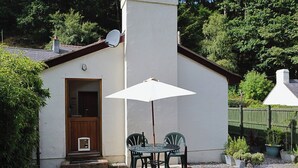 A small, white building with a door and a satellite dish. There is a table with two chairs and a white umbrella. The building is surrounded by trees and gravel. - 1 bedroom accommodation in Capelulo, near Conwy (Capelulo, near Conwy)