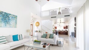 A spacious living area with a white sofa, a glass coffee table, and a dining area in the background.