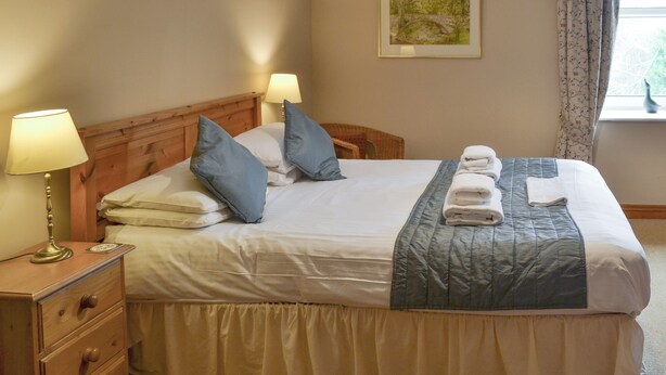 Cama bem arrumada com almofadas, abajur, mesa de cabeceira de madeira e janela com cortinas.