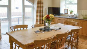 A dining area with a wooden table and chairs, a vase with flowers, and a television in the background.