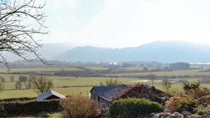 Ländliche Landschaft mit einem Steinhaus, umgeben von Feldern und Bergen im Hintergrund.