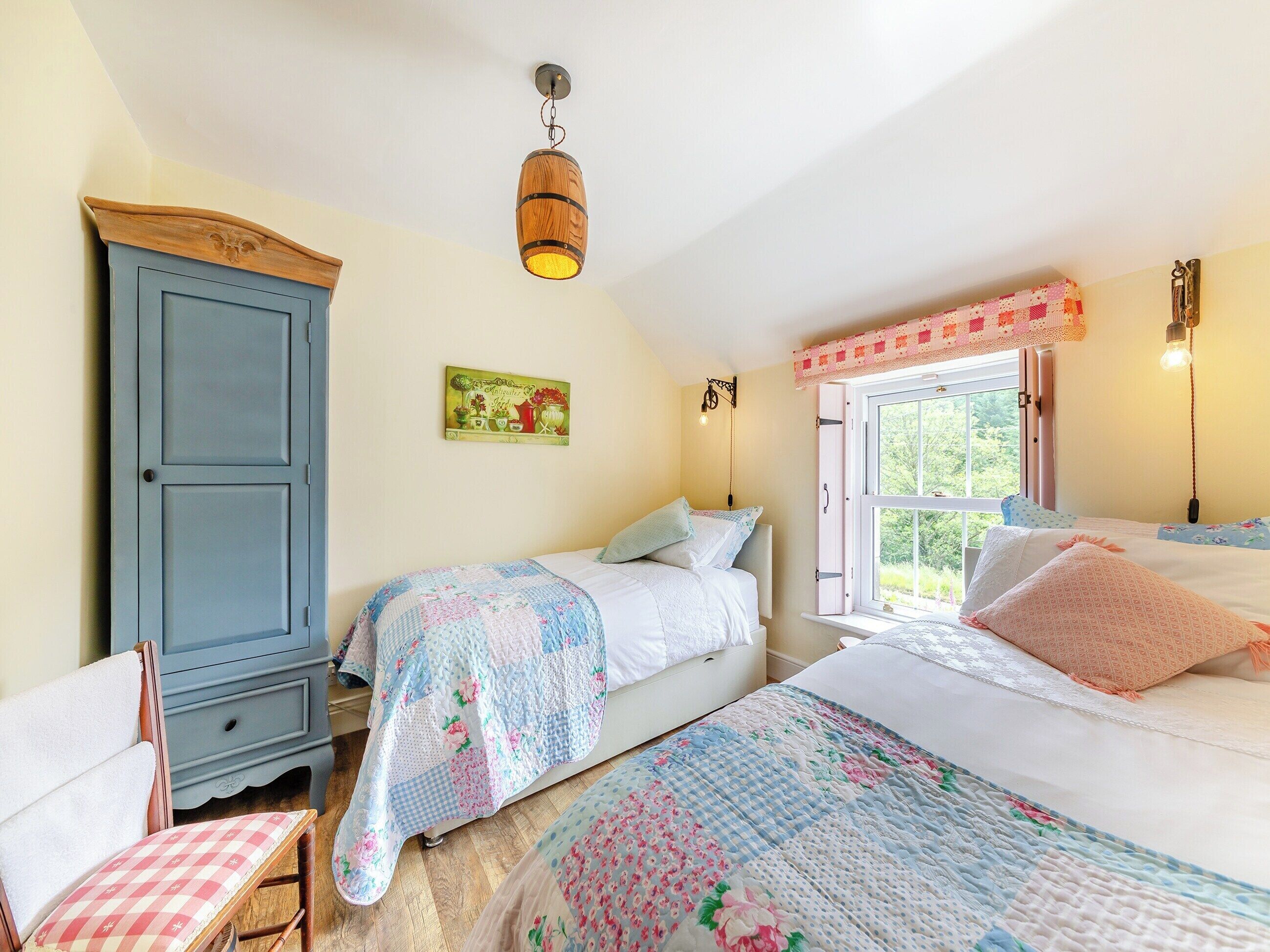 A bedroom with two beds, a wooden wardrobe, a chair, a window with a view, and a wall-mounted light fixture.