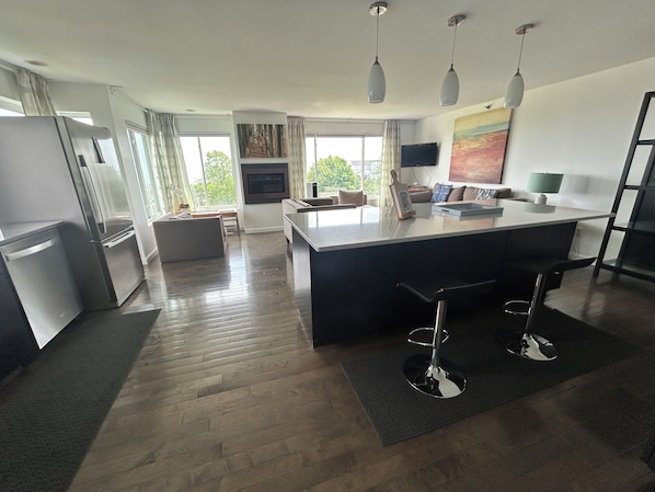A modern kitchen with a central island, bar stools, a dining area with a sofa, and a large window with curtains.
