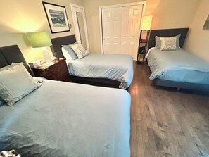Habitación de hotel con tres camas, piso de madera, mesita de noche con lámpara, un cuadro enmarcado en la pared y una puerta.