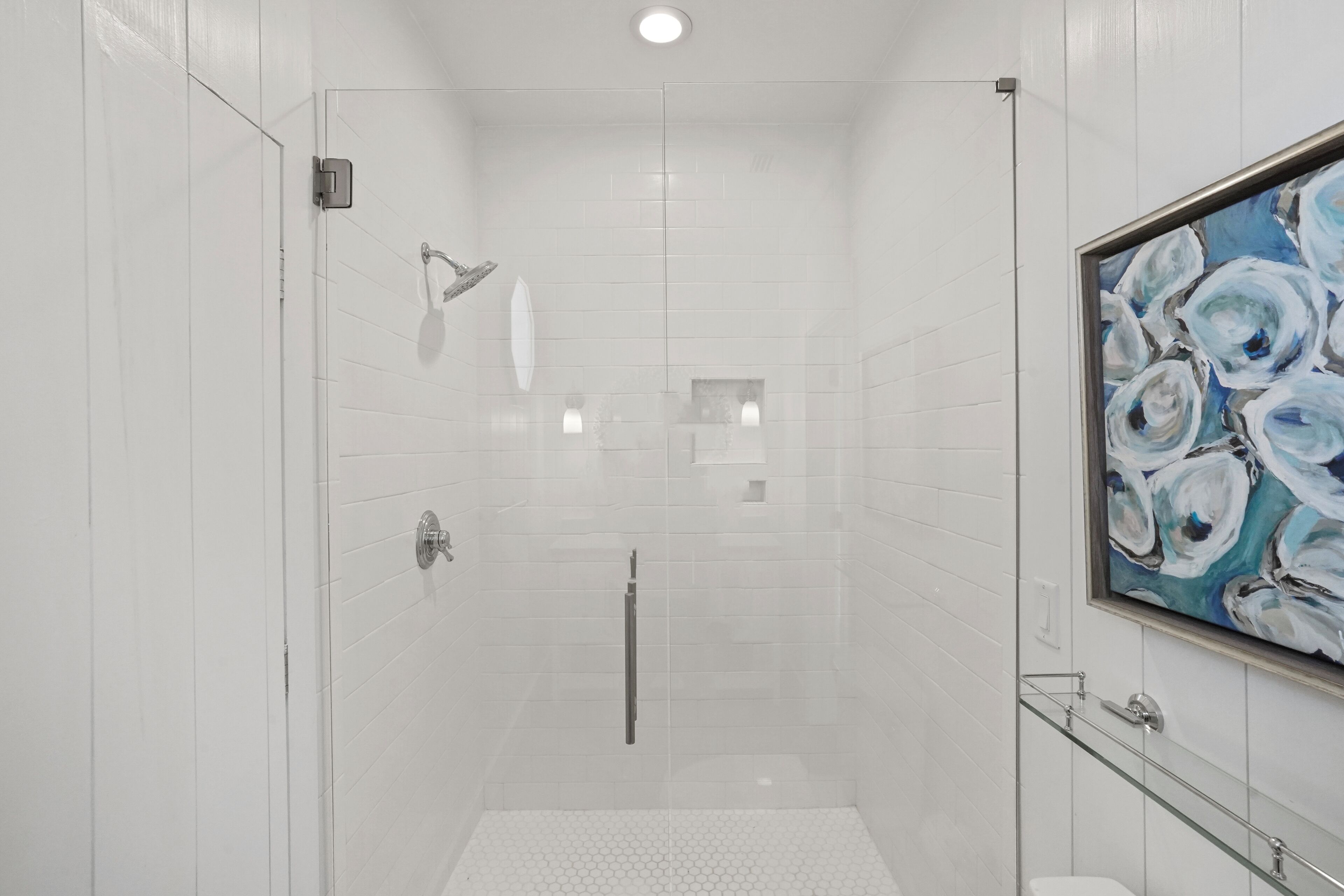 A modern bathroom with white tiles, a glass shower enclosure, a toilet, and a framed painting on the wall.
