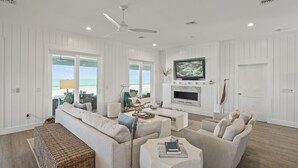 A spacious living room with a large TV, fireplace, and a view of the ocean through the windows. - Graceland: Stunning Views, Screened Porch, End of North Cape! (Cape San Blas)