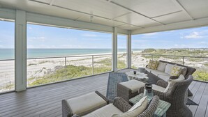 A covered outdoor seating area with wicker furniture, a view of the beach, and a clear sky. - Graceland: Stunning Views, Screened Porch, End of North Cape! (Cape San Blas)
