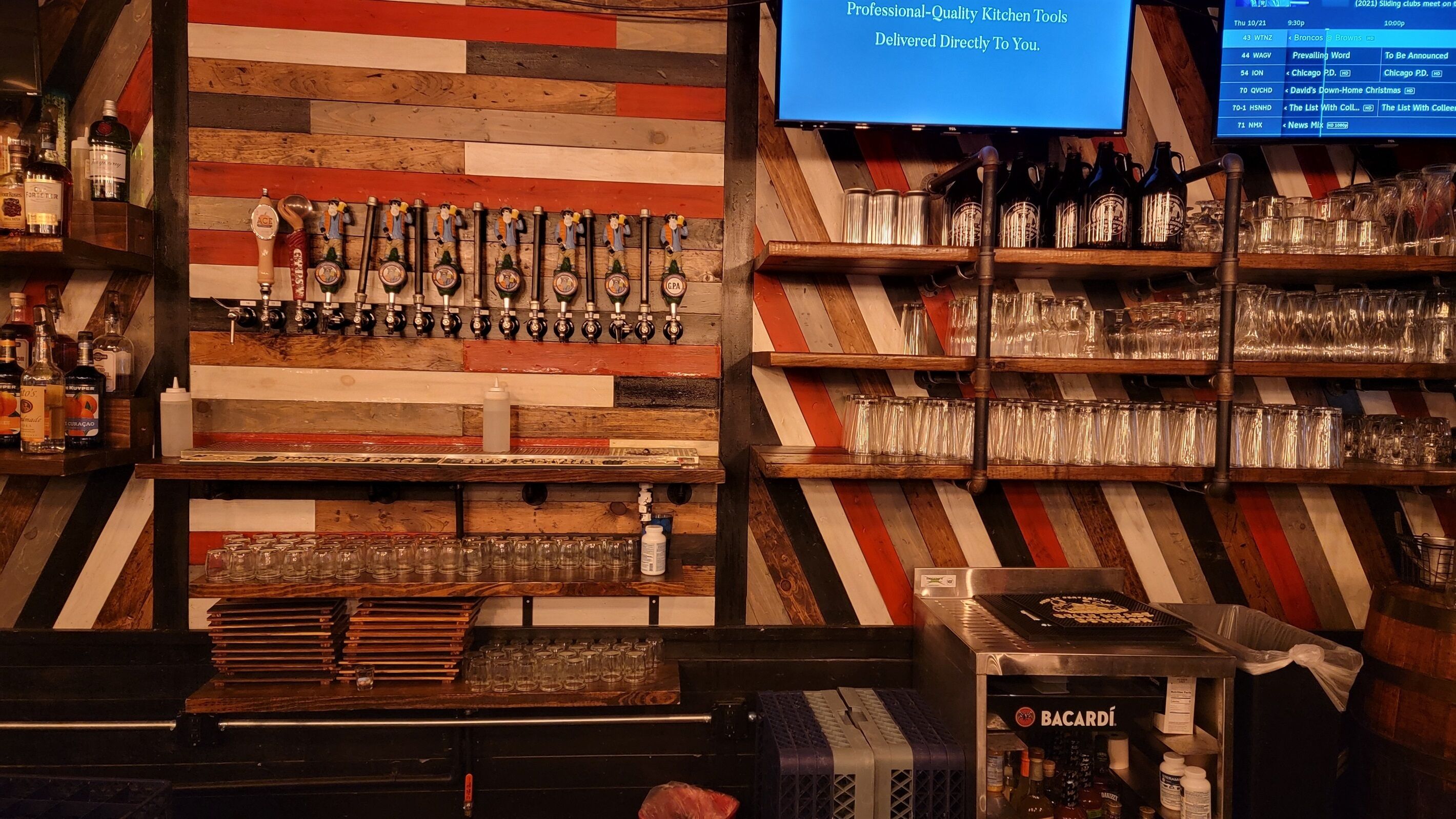 A bar area with a wooden wall, multiple taps, shelves with glasses, and a Bacardi cooler.