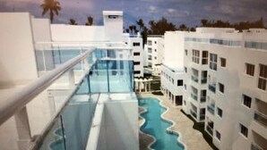 A pool area with a wavy edge, surrounded by white buildings with multiple floors and balconies. - Beautiful Punta Cana Beach Escape! (El Cortecito)