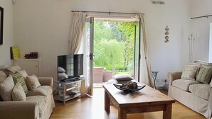 A living room with two sofas, a wooden coffee table, a television on a stand, and a view of a garden through an open door. - Wee Bridge Farm Cottage (Mobberley, near Knutsford)
