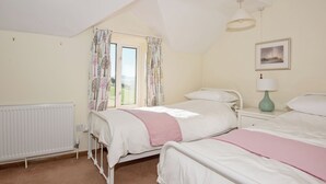 A bedroom with two beds, a window with curtains, a radiator, a bedside table with a lamp, and a framed picture on the wall.